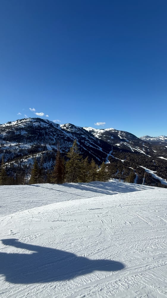 A scenic winter landscape with snow-covered mountains with 3 connected peaks with ski runs taken from across a valley with a groomed ski slope in the foreground.