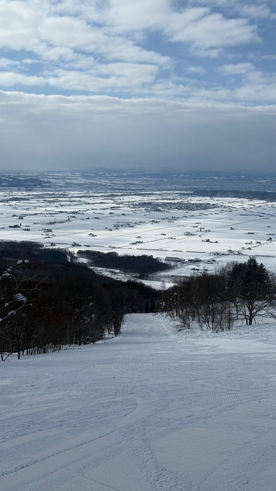 A snowy mountain landscape with a winding ski trail in the foreground and a cloudy sky in the background.