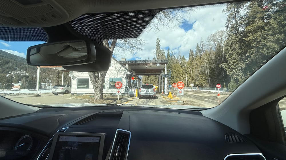 The image shows the view from inside a car driving towards the small Canadian border patrol building. The toll booth is surrounded by trees and mountains, with stop signs and other traffic control signs visible.