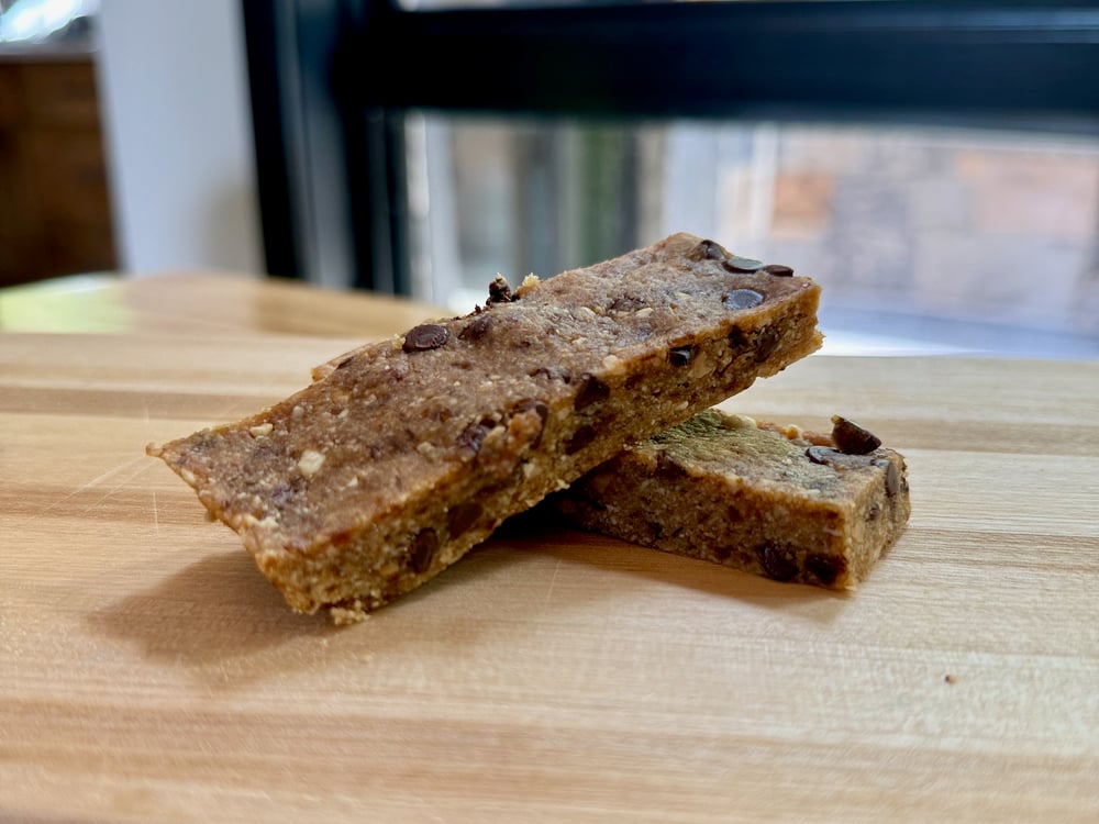 The image shows two rectangular-shaped snack bars or energy bars on a wooden surface. The bars have a brown, nutty-looking appearance with visible chocolate chips embedded in them.