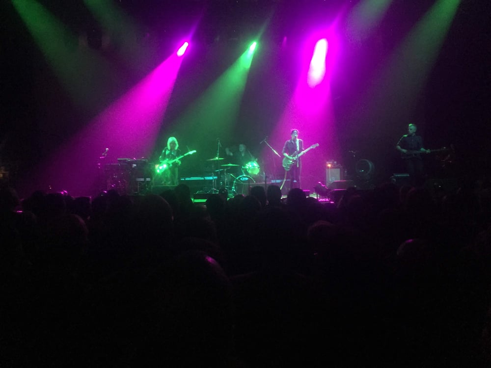 The image shows a live music performance on stage, with the band members playing their instruments under colorful stage lighting. The stage is illuminated by bright pink, green, and purple spotlights, creating a vibrant and energetic atmosphere.