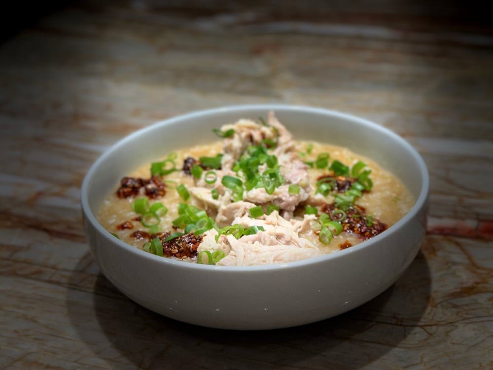 A white bowl on a marble counter. The bowl contains a rice porridge, topped with green onions and shred chicken, drizzled with a dark red chili crunch topping.
