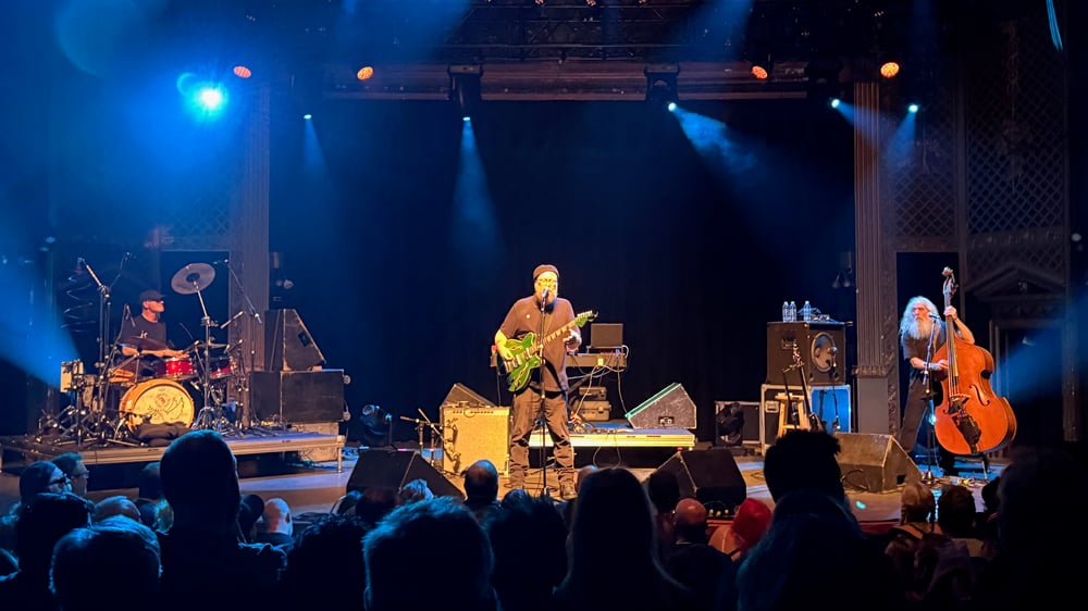 A live music performance on stage with a band playing various instruments, including drums, guitar, and cello, under colorful stage lighting.