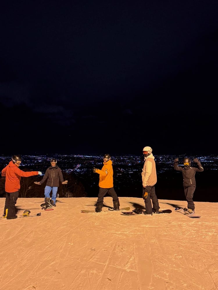 A group of people standing on a snowy slope at night, with the city lights visible in the background.