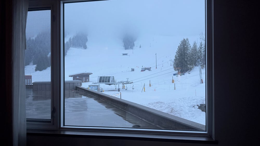 A view of a snowy ski resort through a window, showing ski lifts, slopes, and a foggy winter landscape.