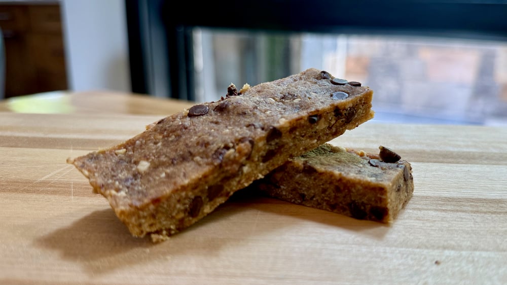 The image shows two pieces of a homemade snack bar or energy bar on a wooden surface. The bars are made with dates, nuts, and chocolate chips, and have a crumbly, textured appearance.