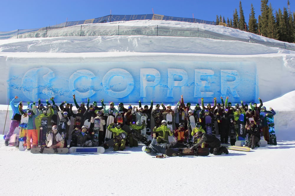 A large group of snowboarders posing in front of a large snow sculpture that spells out 'COPPER' in front of the competition half-pipe