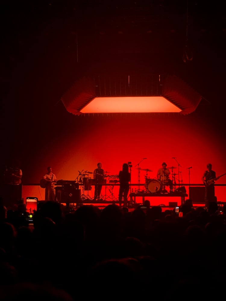 A live music performance on stage with a band playing instruments under a large, glowing orange light fixture.