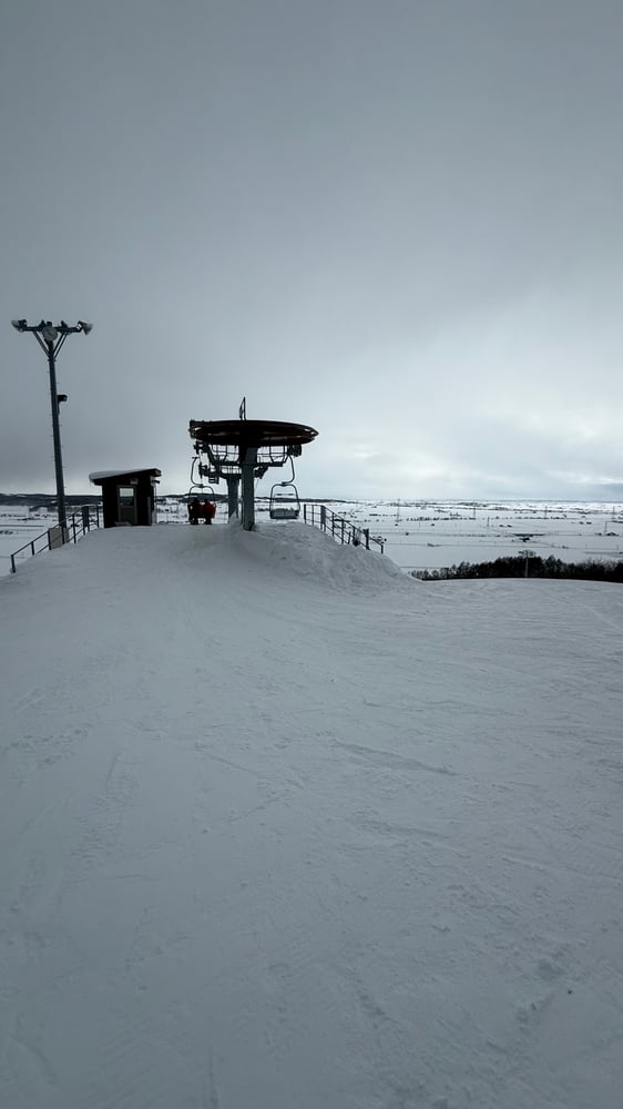A snow-covered ski slope with a chairlift in the distance, set against a cloudy winter landscape.