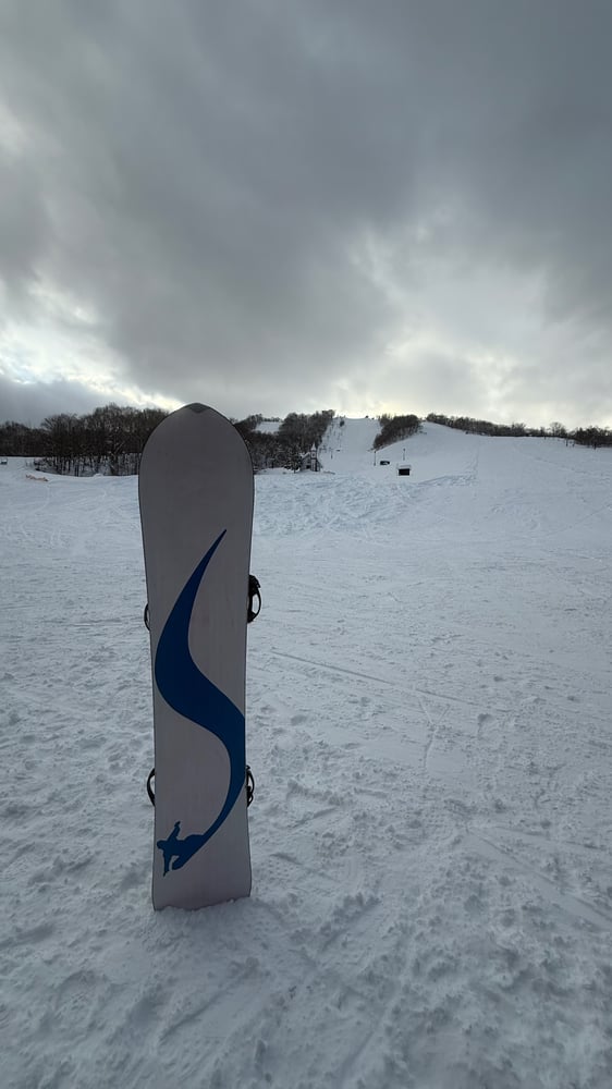 A snowy ski slope with a snowboard standing upright in the foreground on a cloudy winter day.