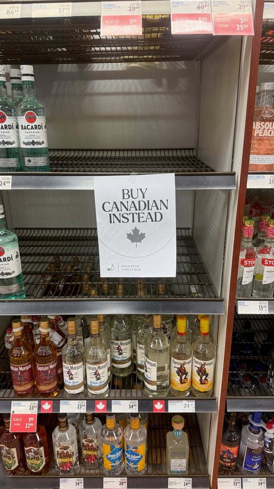 The image shows shelves in a liquor store, with various alcoholic beverages such as Bacardi rum and Captain Morgan rum. A sign on an empty shelf in the center reads 'Buy Canadian Instead', promoting the purchase of Canadian products.
