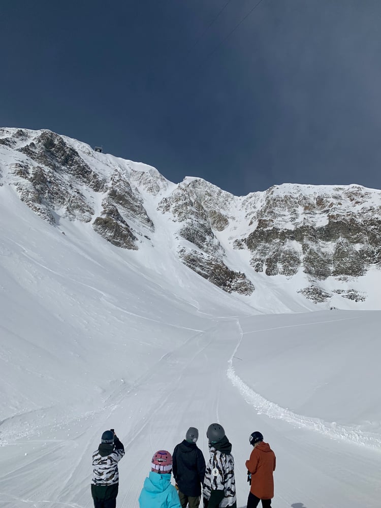 The image shows a group of skiers standing on a snowy mountain slope in Big Sky, MT, looking up at the Big Couloir, a narrow path of snow between large cliffs.