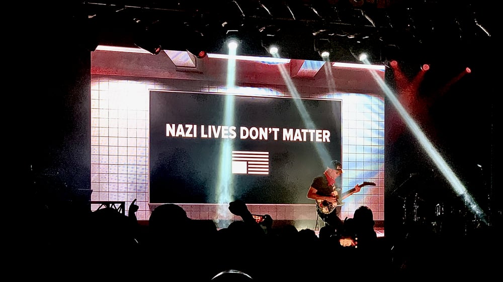 The image shows a large stage with a projection screen displaying the text 'NAZI LIVES DON'T MATTER'. The stage is dimly lit, with spotlights shining on the screen. A guitarist plays in front of the screen. There is a crowd of people in the foreground, watching the performance.
