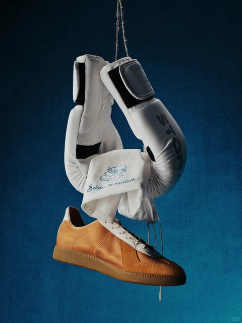 This image presents a highly detailed, photorealistic still-life composition with a studio-quality rendering technique, designed as a style reference for a moody, tactile aesthetic. The dominant color language is a sophisticated contrast between cool and warm tones: a deep, desaturated teal-blue background with a subtle gradient and fine grain texture provides a cool, atmospheric base, while the warm, honeyed tan of the leather sneaker and the crisp, clean white of the boxing gloves and cloth create a striking, focused color story. The lighting is directional and soft, emanating from the upper left to cast gentle, diffused shadows that define the forms and enhance the three-dimensionality of the objects, creating a contemplative and slightly dramatic mood. Texture and material feel are rendered with exceptional clarity; the buttery, fine-grained leather of the sneaker, the smooth, matte synthetic surface of the boxing gloves, the soft, woven cotton of the cloth, and the coarse, fibrous twine are all distinctly tactile. The composition is a centered, vertical arrangement where the objects are suspended and intertwined in a gravity-defying, sculptural manner, creating a shallow but defined depth of field against the flat, textured backdrop. Shape language combines the soft, organic curves of the gloves and shoe with the sharp, geometric lines of the glove straps and stitching, all rendered with precise, clean edges. The overall mood is one of quiet intensity, artistic juxtaposition, and crafted elegance, with a visual weight centered on the interplay of textures and the suspended, dynamic arrangement of the objects.