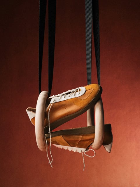 This image presents a photorealistic studio photograph with a highly stylized, commercial aesthetic, characterized by a warm, monochromatic color language dominated by deep terracotta and burnt sienna tones in the background, contrasted by the tan, cream, and brown hues of the leather sneakers. The lighting is soft and directional, emanating from the upper left to create gentle gradients and subtle shadows that emphasize the three-dimensional form of the shoes and the texture of the materials, conveying a mood of sophisticated minimalism and tactile luxury. The composition is centered and vertically oriented, featuring the sneakers suspended by their black laces against a seamless, textured backdrop, creating a sense of balanced asymmetry and focused depth. The texture and material rendering are exceptionally detailed, highlighting the smooth grain of the leather, the soft nap of the suede panels, and the matte finish of the rubber soles, while the shape language blends the organic curves of the footwear with the strict geometric lines of the hanging laces. The overall atmosphere is calm, curated, and premium, evoking a sense of refined craftsmanship and timeless style.