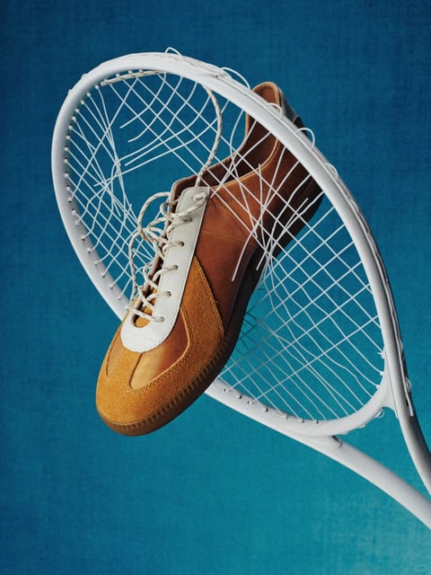 The image presents a photorealistic still life composition with a clean, commercial studio aesthetic. The color language is defined by a high-contrast, complementary palette, pairing the cool, saturated teal of the textured background with the warm, earthy tones of the shoe—rich amber, burnt orange, and chocolate brown suede and leather—accented by the crisp white of the shoe's tongue and the lacrosse stick. Lighting is directional and relatively hard, coming from the upper left to cast defined shadows within the netting and beneath the shoe, creating a sense of three-dimensional form and highlighting the tactile material qualities. The texture and material feel are rendered with high fidelity: the fuzzy nap of the suede, the smooth grain of the leather, the taut, woven nylon of the net, and the matte plastic of the stick frame are all distinctly palpable. Compositionally, the image uses a dynamic diagonal arrangement, with the lacrosse stick's head framing the shoe at an angle, creating depth through the layering of the net over the footwear. Shape language contrasts the organic, curved silhouette of the shoe with the rigid, geometric oval of the stick head and the linear grid of the net. The mood is one of focused, energetic stillness, evoking a sense of athletic preparation or a paused moment in action, with a strong visual weight anchored by the centrally placed, detailed objects against the expansive, simple background.