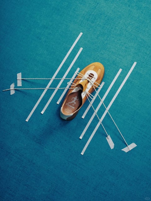 This image presents a stylized, flat-lay composition with a distinct graphic and tactile aesthetic, blending photorealistic object rendering with illustrative, handcrafted elements. The dominant color language is a vibrant, saturated teal blue background that contrasts sharply with the warm mustard-yellow and white of the central shoe, creating a cool-warm dynamic with high contrast. The lighting is soft and diffused, appearing to come from an overhead source, casting gentle, subtle shadows that give the objects a slight lift from the surface without creating harsh drama. The texture and material feel are pronounced: the background has a fine, paper-like grain, the shoe displays soft suede and smooth leather textures, and the paper strips and masking tape have a matte, fibrous quality. Compositionally, the arrangement is dynamic and asymmetrical, using diagonal lines of paper strips and taut strings to create directional energy and a sense of suspended motion around the centrally placed shoe. The shape language combines the organic, curved form of the footwear with the strict, geometric lines of the paper strips and the sharp angles of the tape, creating a visual tension between soft and hard edges. The overall mood is playful, creative, and meticulously arranged, evoking a sense of deconstructed motion or a paused moment in a crafting process.