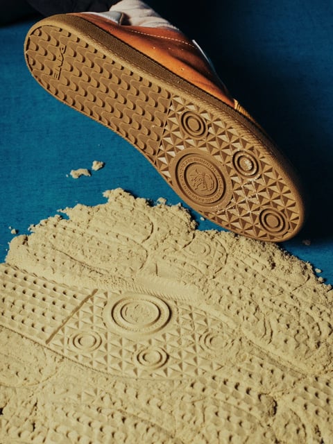 The image presents a photorealistic, high-detail close-up with a shallow depth of field, focusing sharply on the sole of a brown leather shoe and its impression in fine, beige sand. The visual aesthetic is grounded in tactile realism, emphasizing material textures with a crisp, documentary-like clarity. The color language is dominated by a warm, earthy palette: rich amber and cognac tones of the shoe's leather and rubber sole contrast against the cool, deep teal-blue background surface and the neutral, sandy beige of the granular material. Lighting is directional and slightly hard, coming from the upper left, casting defined shadows that accentuate the three-dimensional texture of the sole's tread pattern and the granular structure of the sand, creating a mood of focused, tangible detail. The texture and material feel are paramount: the rubber sole exhibits a matte, slightly worn finish with precise geometric treads, while the sand appears fine, dry, and loosely packed, capturing the impression with high fidelity. Compositionally, the framing is tight and diagonal, with the shoe entering from the upper left, its sole hovering just above the sand, which occupies the lower two-thirds, creating a dynamic spatial relationship and emphasizing the action of imprinting. Shape language combines the organic, irregular contours of the sand pile with the strict geometric patterns of the sole's tread—circles, triangles, and repeating semi-circles—highlighting a contrast between natural and manufactured forms. The overall mood is one of quiet, deliberate action and material exploration, with a visual weight that feels grounded and texturally immersive.