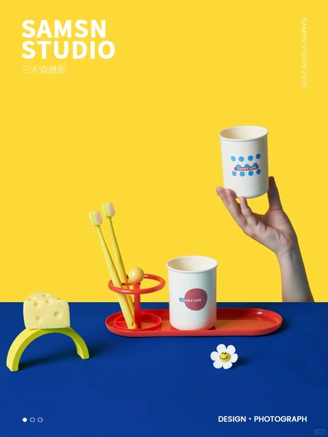 This image is a high-quality, photorealistic still life photograph with a distinct, minimalist, and playful aesthetic, characterized by a clean, graphic composition. The color language is dominated by a vibrant, high-saturation primary palette, featuring a bold, flat yellow background and a deep, solid blue foreground surface, creating a strong, high-contrast division. The objects introduce accents of bright orange, soft white, and pale yellow, maintaining a cohesive, cheerful, and modern color temperature. Lighting is directional and hard, coming from the upper left, casting sharp, well-defined shadows that enhance the three-dimensionality of the objects and create a crisp, studio-quality look. The texture and material feel are rendered with smooth, matte surfaces on the plastic or silicone objects (the toothbrush holder, tray, and cups) and a soft, porous texture on the cheese-like sponge, all appearing clean and tactile. The composition is meticulously arranged with a balanced, asymmetrical layout, utilizing negative space effectively and placing objects along clear horizontal and vertical axes, with a shallow depth of field that keeps the foreground elements sharp. Shape language is predominantly geometric and rounded, featuring simple, bold forms like circles, cylinders, and arches with clean, defined edges and minimal line weight. The overall mood and atmosphere are bright, optimistic, and energetic, conveying a sense of organized, contemporary design with a touch of whimsy.