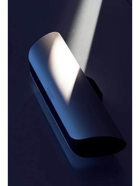 The image presents a photorealistic 3D rendering of a sleek, minimalist electronic device, likely a portable speaker or power bank, with a smooth, matte-finished surface. The color language is dominated by a cool, deep blue-grey palette, with the object's surface reflecting a soft, diffused highlight that transitions from a bright, cool white to a subtle, warm amber tone, creating a striking contrast against the deep, almost black shadows. The lighting approach utilizes a single, strong directional light source from the upper left, casting a hard-edged, dramatic shadow that emphasizes the object's form and creates a high-contrast, moody atmosphere. The texture and material feel are rendered with a fine, uniform grain, suggesting a high-quality plastic or anodized metal with a soft-touch finish, while the sharp, clean edges and seamless construction convey precision engineering. The composition is a close-up, diagonal arrangement that fills the frame, using a shallow depth of field to keep the focus on the illuminated top surface, with the background receding into a deep, uniform blue-grey void. The shape language is a blend of soft, organic curves on the top surface and a sharp, geometric, elongated oval profile, with perfectly smooth, rounded edges. The overall mood and atmosphere are sophisticated, modern, and slightly mysterious, with a calm, high-tech elegance conveyed through the controlled lighting and minimalist design.