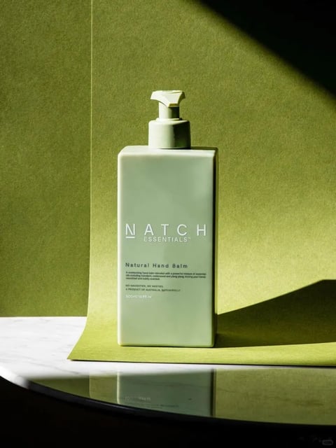 This image presents a photorealistic, minimalist product photograph with a clean, contemporary aesthetic. The dominant color language is a monochromatic, desaturated sage green palette, applied to both the product bottle and the textured paper backdrop, creating a cohesive, tonal look with subtle variations in shade rather than hue. The lighting is directional and hard, coming from the upper right, casting a sharp, defined shadow of the bottle onto the backdrop and creating a strong contrast between the brightly lit surfaces and the deep, dark shadows. The texture and material feel are rendered with high fidelity: the matte, slightly granular surface of the paper backdrop contrasts with the smooth, semi-matte plastic of the bottle and the glossy, reflective marble surface below. Compositionally, the image uses a centered, vertical framing with a slight low-angle perspective, placing the bottle on a reflective surface that creates a mirrored reflection, adding depth. The shape language is predominantly geometric, characterized by the rectangular bottle with sharp edges and the clean, straight lines of the backdrop and surface, contrasted by the organic curve of the pump dispenser. The overall mood is calm, sophisticated, and clinical, with a high visual weight due to the strong contrast and central subject placement.