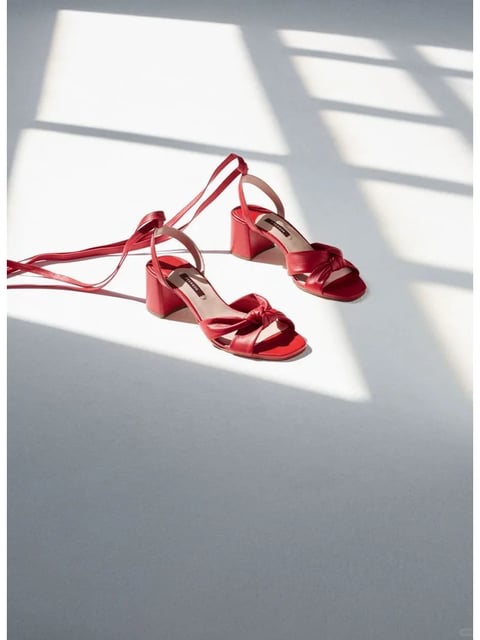 This image presents a photorealistic, high-fashion style reference with a clean, minimalist aesthetic. The dominant color language features a saturated, vibrant cherry red against a cool, neutral light gray background, creating a striking contrast with high saturation on the subject and low saturation in the negative space. The lighting approach utilizes strong, directional natural light, likely from a window, casting sharp, geometric shadows that create a high-contrast interplay of light and dark across the scene. The texture and material feel are rendered with precision, highlighting the smooth, semi-glossy finish of the red leather, the subtle grain of the insole, and the crisp, hard edges of the block heels. Compositionally, the image employs an asymmetrical, diagonal arrangement with the shoes placed off-center, using the dramatic shadows to create depth and lead the eye. The shape language combines the organic curves of the knotted straps and footbeds with the rigid, geometric forms of the block heels and the sharp, rectangular shadow patterns. The overall mood and atmosphere convey a sense of modern elegance, sophisticated minimalism, and a calm, sun-drenched stillness.