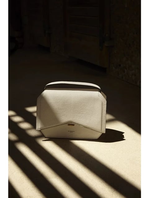 This is a photorealistic product photograph with a minimalist, high-fashion editorial aesthetic. The image features a single, structured handbag in a creamy off-white color, rendered with a fine, pebbled texture that suggests a high-quality leather or exotic skin material. The color palette is dominated by warm, neutral tones—creamy whites, soft beiges, and deep, warm shadows—with low saturation and high contrast between the brightly lit foreground and the dark, shadowed background. The lighting is dramatic and directional, coming from a low angle to the left, casting long, sharp, diagonal shadows across the smooth, light-colored floor, creating a strong sense of depth and a serene, almost contemplative mood. The composition is centered and balanced, with the bag placed as the sole subject in a sparse, architectural space, framed by the interplay of light and shadow. The shape language is a mix of clean, geometric lines from the bag's structure and the sharp shadows, contrasted with the soft, organic texture of its surface. The overall atmosphere is one of quiet luxury, sophistication, and focused attention on material and form.