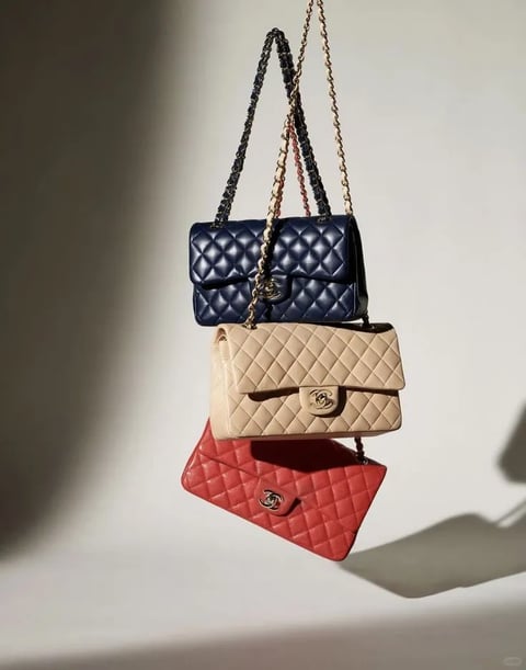 The image presents a photorealistic, high-fashion still life with a clean, minimalist aesthetic. The dominant color language features a sophisticated trio of deep navy blue, soft beige, and vibrant coral red, all rendered with rich saturation and strong contrast against a neutral, light gray background. The lighting is directional and hard, casting sharp, defined shadows to the right, creating a dramatic, high-contrast mood with a clear sense of volume. The texture and material feel are meticulously rendered, showcasing the supple, quilted leather with a subtle sheen and the polished, reflective quality of the gold-tone chain straps and interlocking 'CC' clasps, conveying a luxurious tactile impression. The composition is vertically stacked and centered, with the three bags suspended in mid-air to create a dynamic, gravity-defying arrangement that emphasizes depth through overlapping forms and cast shadows. The shape language is defined by the structured, geometric rectangular forms of the bags, softened by the rounded edges of the quilting pattern and the fluid, organic drape of the chain straps. The overall mood is elegant, aspirational, and meticulously styled, with a high visual weight that conveys luxury and precision.
