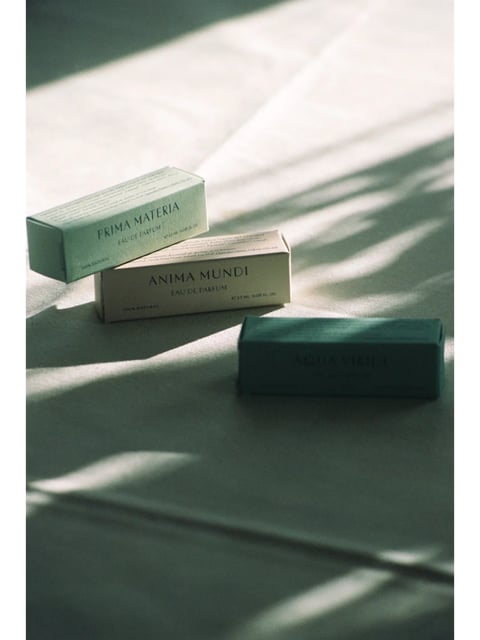 The image presents a photorealistic still life of three small, rectangular perfume sample boxes arranged on a textured, light gray fabric surface, likely linen or cotton, under strong directional lighting. The visual aesthetic is clean, minimalist, and tactile, emphasizing natural materials and subtle details. The color language is muted and earthy, dominated by a palette of sage green, warm beige, and deep teal, with low saturation and soft contrast that enhances the natural, organic feel. The lighting approach is characterized by hard, directional sunlight casting long, sharp shadows from an unseen window frame or blinds, creating a dramatic interplay of light and shadow that adds depth and a serene, contemplative mood. Texture and material feel are prominent, with the matte, slightly fibrous paper of the boxes and the woven texture of the fabric surface rendered with high detail, inviting a tactile impression. Compositionally, the image uses an asymmetrical, diagonal arrangement of the boxes, with a shallow depth of field that keeps the foreground box in sharp focus while softly blurring the background, creating a sense of intimacy and focus. Shape language is geometric, with the clean lines and right angles of the boxes contrasting against the organic, irregular folds of the fabric. The overall mood and atmosphere are calm, sophisticated, and introspective, evoking a sense of quiet luxury and natural simplicity.