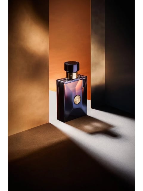 This image is a high-end, photorealistic product photograph with a sophisticated, luxurious aesthetic. The dominant color language features a warm, amber-to-terracotta background contrasted with deep, cool-toned shadows in indigo and black, creating a high-contrast, dramatic palette with rich saturation. The lighting approach is directional and hard, originating from the upper left, casting sharp, elongated shadows and creating bright, specular highlights on the glass bottle, which conveys a mood of elegance and mystery. The texture and material feel are meticulously rendered, showcasing the smooth, reflective, and faceted dark glass of the bottle, the metallic sheen of the gold cap and emblem, and the matte, slightly granular texture of the paper-like background surfaces. The composition is asymmetrical and architectural, with the bottle positioned off-center within a geometric arrangement of vertical and horizontal planes that create a strong sense of depth and structured space. The shape language is predominantly geometric, defined by the bottle's sharp, angular facets, the cylindrical cap, and the clean, straight lines of the background panels, with precise edge treatment. The overall mood and atmosphere are one of refined luxury, quiet intensity, and modern sophistication, with a heavy visual weight anchored by the dark bottle and its pronounced shadow.