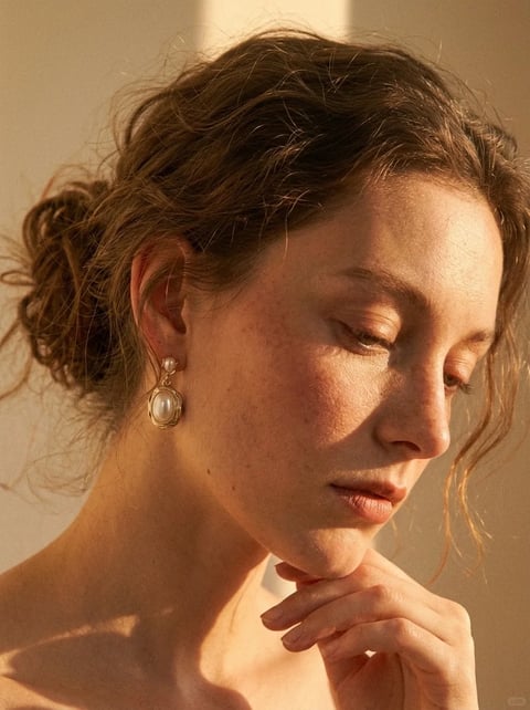 This image is a photorealistic portrait with a painterly, fine-art quality, rendered with exceptional detail that captures the subtle textures of skin, hair, and fabric. The color language is dominated by a warm, monochromatic palette of soft amber, golden ochre, and muted peach tones, with low saturation and gentle contrast that creates a cohesive, sun-drenched feel. The lighting approach features soft, directional light coming from the upper left, casting delicate, diffused shadows that model the face and neck with a gentle, naturalistic quality, evoking a serene, contemplative mood. Texture and material feel are rendered with high fidelity, showcasing the fine grain of skin, the wispy, flyaway strands of hair, and the smooth, luminous surface of the pearl earring, creating a tactile impression of organic softness and delicate refinement. The composition is a tight, intimate close-up, framing the subject's face and hand in a three-quarter view with a shallow depth of field that softly blurs the background, focusing attention on the subject's features and gesture. Shape language is predominantly organic, defined by the soft curves of the face, the loose, flowing lines of the hair, and the gentle contours of the hand, with soft edge treatment that enhances the image's gentle, naturalistic feel. The mood and atmosphere are deeply serene, introspective, and quietly elegant, with a low-energy, contemplative visual weight that emphasizes stillness and subtle beauty.