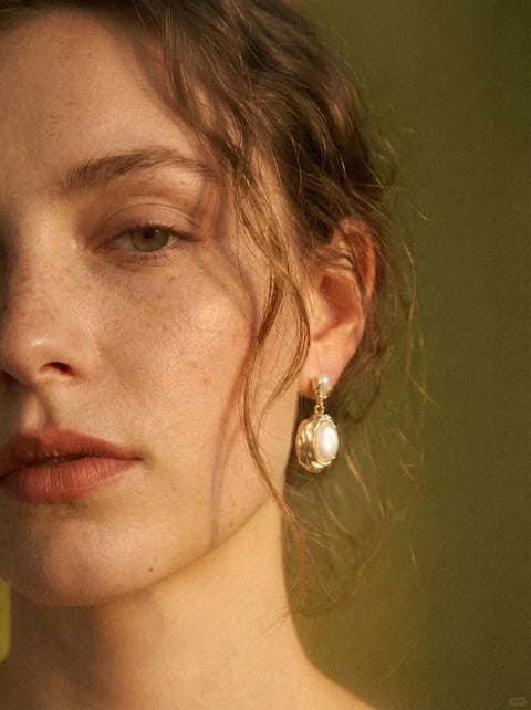This image presents a photorealistic, painterly portrait with a warm, vintage aesthetic. The color language is dominated by a muted, earthy palette of soft olive green, warm ochre, and desaturated peach tones, with low saturation and gentle contrast that creates a harmonious, tonal quality. The lighting is soft and directional, appearing to come from the upper left, casting gentle, diffused shadows that sculpt the face with a natural, intimate glow. The texture and material feel are highly tactile, emphasizing the realistic grain of the skin, the fine, wispy strands of curly hair, and the smooth, lustrous surface of the pearl earring set in textured gold. Compositionally, it is a tight, asymmetrical close-up, cropping the face to focus on the eye, cheek, and ear, creating a shallow depth of field that softly blurs the background. The shape language is organic, defined by the natural curves of the facial features, the soft waves of hair, and the rounded form of the jewelry, with soft edge treatment. The mood and atmosphere are serene, contemplative, and quietly elegant, conveying a sense of timeless, natural beauty with low visual energy.
