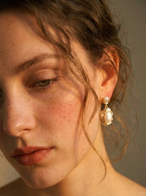 The image presents a photorealistic, painterly style reference with a warm, intimate aesthetic. The color language is dominated by a warm, earthy palette of peachy skin tones, soft terracotta lips, and deep auburn hair, accented by the creamy white of the pearl earring and the subtle gold of its setting, all rendered with a muted, low-saturation quality and soft contrast. The lighting approach is directional and soft, coming from the upper left, casting gentle, diffused shadows that sculpt the face and create a contemplative, quiet mood. The texture and material feel are highly tactile, emphasizing the natural, slightly dewy skin with visible freckles and pores, the fine, wavy strands of damp-looking hair, and the smooth, luminous surface of the pearl against the textured gold metal. The composition is a tight, intimate close-up, framing the right side of the face and ear with a shallow depth of field that keeps the focus sharp on the skin and earring while softly blurring the background. The shape language is predominantly organic, defined by the soft curves of the facial features, the ear, and the loose, wavy hair, with the earring providing a contained, geometric oval form. The overall mood and atmosphere are serene, introspective, and slightly melancholic, with a low energy level and significant visual weight placed on the subject's downward gaze and the delicate details of the skin and jewelry.