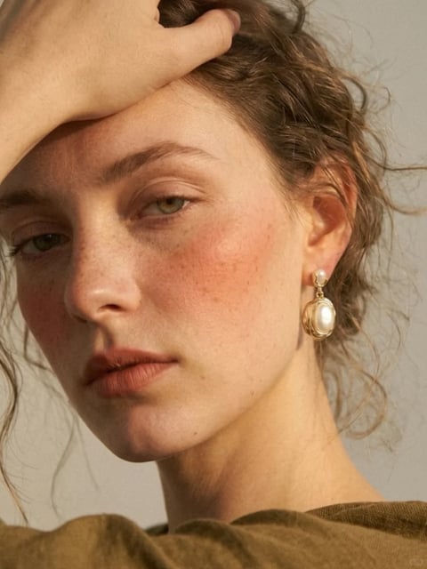 This is a photorealistic, close-up portrait rendered with a soft, painterly quality, emphasizing a natural and intimate aesthetic. The color language is dominated by warm, earthy tones: the subject's skin has a natural, slightly flushed complexion with subtle freckles, complemented by the muted olive-green of her top and the soft, neutral beige background. Saturation is moderate and natural, with contrast that is gentle yet defined, particularly in the soft shadows around the eyes and under the chin. The lighting approach is soft and directional, appearing to come from the upper left, casting a gentle glow on the forehead and cheek while creating soft, diffused shadows that enhance the three-dimensional form without harshness. Texture and material feel are highly detailed and tactile: the skin shows fine pores and natural imperfections, the hair has a wispy, slightly messy texture with individual strands catching the light, and the gold and pearl earring has a smooth, reflective surface with a subtle metallic sheen. Compositionally, this is a tight, intimate framing that crops closely on the face and neck, with the subject's hand resting on her forehead to create a diagonal line that draws the eye inward, emphasizing depth through shallow focus on the foreground features. Shape language is predominantly organic, defined by the soft curves of the face, the wavy lines of the hair, and the rounded forms of the earring, with edges that are softly blended rather than sharply defined. The overall mood and atmosphere are contemplative and serene, with a quiet, introspective energy conveyed through the subject's direct yet soft gaze and the gentle, warm lighting.