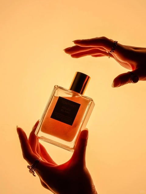 This image is a high-fashion, photorealistic still-life photograph with a luxurious, warm, and sensual aesthetic. The dominant color language is a monochromatic, saturated amber and golden-orange palette, with a strong warm color temperature that bathes the entire scene. The lighting is soft and diffused, appearing to emanate from a large, warm source behind the subject, creating a glowing backlight that silhouettes the hands and bottle while casting gentle, warm highlights on the glass and skin. The texture and material feel are rendered with high fidelity: the glass bottle has a smooth, reflective surface with subtle imperfections and a clear, viscous liquid inside; the metallic cap has a brushed, reflective sheen; and the skin on the hands shows fine detail, including pores and the soft texture of the fingers. The composition is centered and balanced, with the perfume bottle held delicately between two hands in a symmetrical, almost reverent arrangement, creating a shallow depth of field that keeps the bottle in sharp focus against the softly blurred, gradient background. The shape language is a contrast between the sharp, geometric lines of the rectangular bottle and the soft, organic curves of the human hands and fingers. The overall mood is one of elegance, intimacy, and warm, sun-drenched luxury, with a calm and focused energy level.