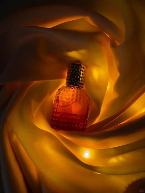 This image is a photorealistic, high-end product photograph with a luxurious, warm aesthetic. The dominant color language is a rich, monochromatic palette of deep amber, honeyed gold, and warm ochre, with high saturation and strong contrast between luminous highlights and deep, velvety shadows. The lighting is dramatic and directional, appearing to emanate from within or behind the central subject, creating a soft, glowing backlight that diffuses through the surrounding material and casts long, soft shadows. The texture and material feel are defined by the contrast between the smooth, faceted glass of the perfume bottle, which refracts light into sharp, geometric patterns, and the soft, flowing, semi-translucent fabric that envelops it, rendered with a silky, liquid-like drape. The composition is centered and immersive, with the bottle nestled in the heart of swirling fabric folds that create a sense of depth and draw the eye inward. The shape language combines the structured, geometric facets of the bottle with the organic, flowing curves of the fabric. The overall mood is one of opulent warmth, intimacy, and sensual elegance, with a heavy visual weight concentrated in the glowing center.