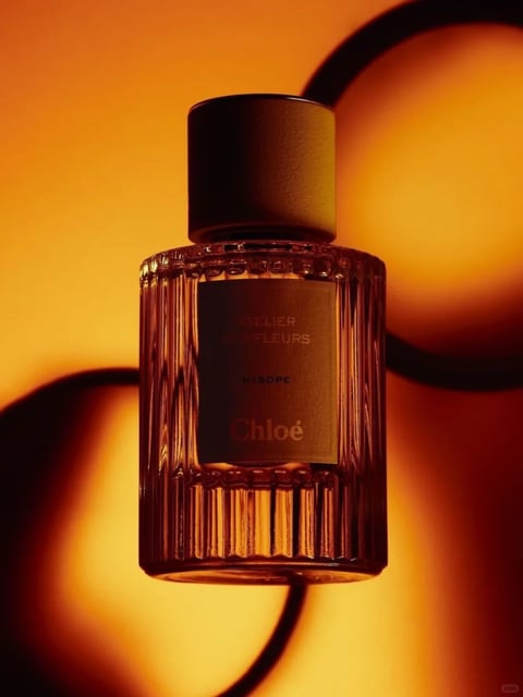 The image presents a photorealistic, high-end product photograph of a Chloé Atelier des Fleurs Hysop perfume bottle, rendered with a luxurious, warm aesthetic. The color language is dominated by a rich, monochromatic palette of deep amber, burnt orange, and honeyed gold, with high saturation and strong contrast created by the interplay of light and shadow. The lighting approach utilizes a strong, directional backlight that creates a glowing, radiant effect, casting the bottle in a warm silhouette with soft, diffused edges and dramatic, elongated shadows that curve across the background. Texture and material feel are emphasized through the crisp rendering of the ribbed, vertical fluting on the glass bottle, which catches the light to create a tactile, crystalline quality, contrasting with the smooth, matte finish of the dark cylindrical cap and the subtle sheen of the metallic label. Compositionally, the bottle is centrally framed in a medium close-up, with a shallow depth of field that blurs the abstract, curvilinear shadow forms in the background, focusing all visual weight on the product. Shape language is a blend of geometric precision in the bottle's cylindrical form and vertical lines, juxtaposed with the organic, flowing curves of the shadow patterns. The overall mood is one of sophisticated warmth, elegance, and intimate luxury, conveyed through the glowing, atmospheric lighting and the rich, enveloping color temperature.