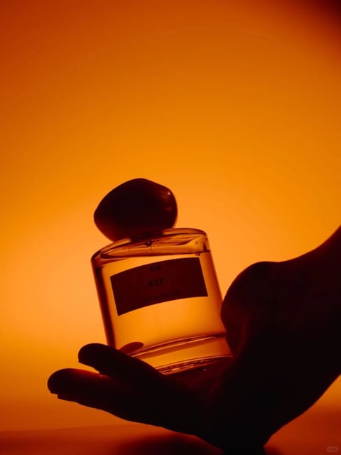 This image presents a photorealistic, high-contrast product photograph with a warm, monochromatic color language dominated by a saturated amber-orange background. The lighting approach is dramatic and directional, creating a strong silhouette effect where the hand and perfume bottle are rendered as deep, dark forms against the luminous backdrop, with soft, diffused light passing through the translucent glass to reveal the liquid inside. The texture and material feel are defined by the smooth, reflective quality of the glass bottle and the matte, organic surface of the stone-like cap, contrasted against the soft, shadowed skin of the hand. Compositionally, the image uses a close-up, centered framing with a shallow depth of field, focusing attention on the bottle held in the palm. The shape language combines the geometric, cylindrical form of the bottle with the organic, irregular shape of the cap and the curved lines of the hand. The overall mood and atmosphere is intimate, luxurious, and warm, evoking a sense of calm and sensory focus through its minimalist yet richly lit presentation.