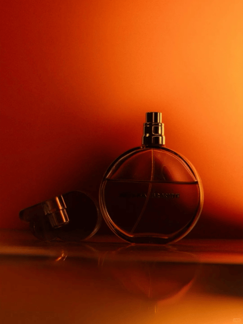 This image presents a photorealistic, product-style still life photograph with a deeply saturated, monochromatic color language dominated by warm amber, burnt orange, and deep sienna tones. The color temperature is intensely warm, with high contrast between the glowing, luminous background and the dark, silhouetted foreground objects. The lighting approach is dramatic and directional, seemingly from behind and to the side, creating strong rim lighting that outlines the contours of the glass bottles and casting deep, soft-edged shadows. The texture and material feel are rendered with high fidelity, emphasizing the smooth, reflective quality of the glass and the polished, metallic sheen of the spray nozzles, with the liquid inside the bottles adding a viscous, translucent depth. The composition is asymmetrical and balanced, featuring a large, upright circular bottle as the primary focal point on the right, with a smaller, tilted bottle lying on its side to the left, all set upon a highly reflective surface that creates a mirrored, blurred reflection below. The shape language is a mix of organic curves from the bottle forms and precise geometric circles and cylinders, with clean, sharp edges defined by the backlighting. The overall mood and atmosphere are luxurious, intimate, and slightly mysterious, evoking a sense of warmth and sophistication through the rich, glowing color palette and the quiet, staged arrangement of the objects.