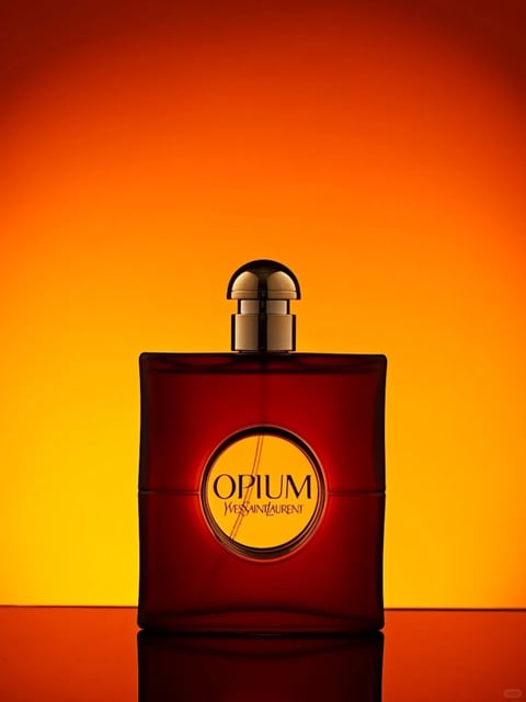The image presents a highly stylized, photorealistic product photograph with a dramatic, cinematic aesthetic. The color language is dominated by a warm, monochromatic palette of deep amber, burnt orange, and rich crimson, with high saturation and strong contrast between the glowing background and the dark, translucent bottle. The lighting is directional and dramatic, creating a strong backlighting effect that makes the bottle's edges and the liquid inside appear to glow with internal warmth, while casting a sharp, dark reflection on the glossy surface below. The texture and material feel are rendered with precision, showcasing the smooth, heavy glass of the bottle, the metallic sheen of the gold cap, and the polished, reflective surface it rests upon. The composition is centered and symmetrical, with the bottle placed prominently in the middle ground against a gradient backdrop that creates a sense of depth and focus. The shape language is a mix of geometric forms—the rectangular bottle and cylindrical cap—contrasted with the organic, circular medallion on the front. The overall mood is luxurious, intense, and mysterious, with a high-energy visual weight conveyed through the bold colors and dramatic lighting.