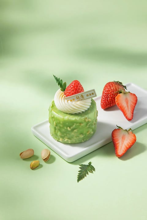 The image presents a photorealistic, high-resolution food photography style with a clean, minimalist aesthetic. The dominant color language features a soft, muted pastel green background that complements the vibrant, saturated reds of the strawberries and the creamy white of the whipped topping, creating a fresh, spring-like palette with gentle contrast and a cool color temperature. Lighting is soft and diffused, appearing to come from the upper left, casting subtle, soft-edged shadows that enhance the three-dimensional form of the dessert without harshness, conveying a calm, inviting mood. Textures are rendered with meticulous detail: the glossy, slightly uneven green coating of the cake reveals embedded nut fragments, the whipped cream shows delicate, swirled ridges, and the strawberries display realistic seed textures and juicy, glistening surfaces, all contributing to a tactile, appetizing feel. Compositionally, the subject is centered on a simple white rectangular plate, with supporting elements like pistachios and strawberry halves arranged asymmetrically in the foreground and background to create depth and visual balance, using a shallow depth of field that keeps the dessert in sharp focus while softly blurring the periphery. Shape language combines the organic, rounded forms of the fruit and cream with the geometric, cylindrical shape of the cake and the rectangular plate, with clean edges and smooth transitions. The overall mood is serene, elegant, and refreshing, evoking a sense of light, natural indulgence.