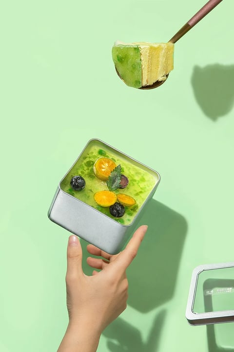 This image exemplifies a clean, modern, and vibrant commercial food photography style, rendered with photorealistic precision. The dominant color language is a fresh, monochromatic palette of soft mint green and lime green, punctuated by high-saturation accents of kumquat orange and deep blueberry purple, creating a cool yet inviting temperature. Lighting is bright and directional from the upper left, casting soft but defined shadows that add dimensionality without harshness, evoking a cheerful, daytime mood. Textures are meticulously rendered: the matte, slightly granular surface of the silver metal tin contrasts with the glossy, translucent gelatinous topping, the delicate, porous crumb of the cake layer, and the fresh, dewy skin of the fruit garnishes. Compositionally, the image uses a dynamic, floating arrangement with the hand-held tin as the central anchor, a spoonful of dessert suspended above, and a partial lid in the lower right, creating depth through layered elements against a flat, seamless background. Shape language balances the geometric, sharp-edged square tin with the organic, irregular forms of the fruit and the soft, rounded fingers. The overall mood is refreshing, appetizing, and stylistically crisp, with a light, airy visual weight that emphasizes the dessert's freshness and appealing texture.