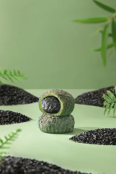 This image presents a photorealistic, studio-style product photograph with a clean, minimalist aesthetic. The dominant color palette is a harmonious blend of soft, muted sage green as the background and surface, contrasted sharply by the deep, matte black of sesame seeds and the vibrant, mossy green of the confectionery. Saturation is moderate, with the greens feeling natural and the blacks rich and dense, creating a high-contrast yet balanced composition. Lighting is soft and diffused, appearing to come from the upper left, casting gentle, subtle shadows that define the forms without harshness, conveying a calm and fresh mood. The texture and material feel are highly tactile: the confectionery has a fine, powdery, almost fuzzy surface coating, while the sesame seeds appear as individual, granular, and slightly glossy specks. The composition is centered and symmetrical, with two stacked, rounded pastries forming the focal point, surrounded by small, distinct mounds of black sesame seeds arranged in a deliberate, almost landscape-like pattern on the flat green plane. Depth is created through a shallow depth of field, with foreground and background elements softly blurred. The shape language is predominantly organic, featuring the soft, irregular spheres of the pastries and the natural, feathery forms of the fern leaves, contrasted by the granular, particulate nature of the seeds. The overall mood is serene, earthy, and sophisticated, with a low-energy, contemplative atmosphere that emphasizes natural ingredients and artisanal craftsmanship.