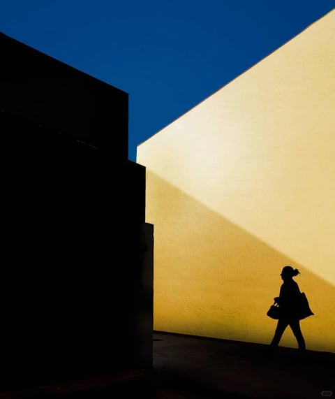 This image presents a striking, high-contrast photographic style defined by bold geometric abstraction and dramatic chiaroscuro. The composition is dominated by a sharp, diagonal division between a deep, saturated cerulean blue sky and a sunlit, warm yellow-ochre wall, creating a powerful color block effect. A large, angular black shadow occupies the left third of the frame, its hard edge cutting cleanly across the scene and emphasizing the stark, graphic quality of the light. The lighting is intense and directional, likely from a low-angle sun, casting a crisp, elongated shadow of a solitary figure walking along the base of the yellow wall. This figure, rendered as a pure black silhouette with a distinct ponytail and carrying a bag, adds a human element and scale to the otherwise minimalist architectural study. The texture of the yellow wall is subtly granular, suggesting stucco or concrete, which contrasts with the smooth, flat appearance of the shadow and sky. The overall mood is one of quiet, cinematic solitude, with a strong emphasis on shape, color field, and the interplay between light and shadow, characteristic of a stylized, graphic photographic approach.
