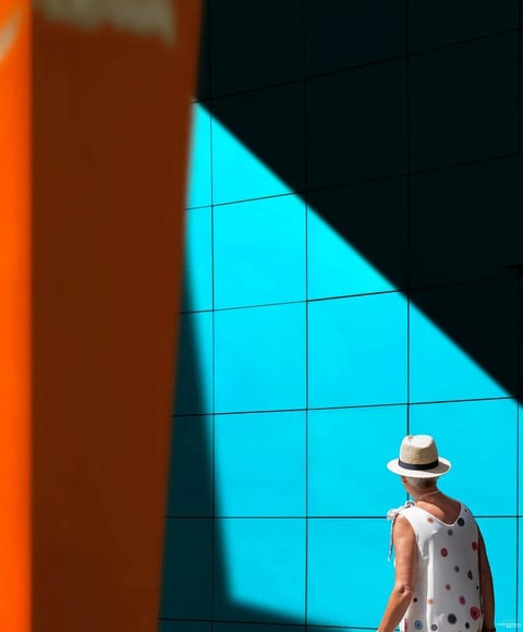 The image is a photorealistic photograph with a strong graphic, minimalist aesthetic, characterized by bold, flat planes of saturated color and sharp geometric divisions. The dominant color palette is a high-contrast, complementary scheme of vibrant cyan-blue and deep, warm orange, with a stark, dark shadow creating a strong diagonal division. The lighting is hard, direct, and directional, casting a crisp, well-defined shadow that emphasizes the geometric shapes and creates a high-contrast, dramatic mood. The texture is smooth and clean, with the tiled wall presenting a flat, matte surface and the foreground orange element appearing as a soft, out-of-focus plane. Compositionally, the image uses a strong diagonal split, with the subject placed off-center in the lower right, creating dynamic tension and depth through the interplay of light, shadow, and the blurred foreground element. The shape language is predominantly geometric, defined by the grid of the wall tiles and the sharp angle of the shadow, contrasted with the organic form of the person. The overall mood is serene yet visually striking, evoking a sense of quiet contemplation within a modern, architecturally defined space.