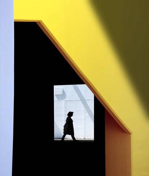The image presents a highly stylized, graphic composition with a strong emphasis on geometric abstraction and bold color blocking. The visual aesthetic is minimalist and architectural, rendered with a flat, almost illustrative quality that prioritizes shape and form over intricate detail. The color language is dominated by a high-contrast palette of saturated primary and secondary colors: a vibrant, warm yellow occupies the upper right, sharply divided by a deep, pure black angular shape, with a sliver of muted blue-gray on the far left and a section of terracotta orange at the lower right. The lighting is dramatic and directional, casting hard, sharp-edged shadows that create a stark division between illuminated and shadowed planes, enhancing the graphic quality. Texture is minimal and smooth, with surfaces appearing as flat, matte planes of color, devoid of tactile detail. The composition is meticulously framed, using architectural elements to create a nested, geometric frame-within-a-frame that focuses attention on a central rectangular opening. Within this opening, a silhouetted figure walks against a brightly lit, pale gray wall, creating a sense of depth and narrative contrast. Shape language is predominantly geometric and angular, with clean, sharp lines and well-defined edges. The overall mood is one of stark, modern elegance and quiet drama, evoking a sense of urban isolation and graphic precision.
