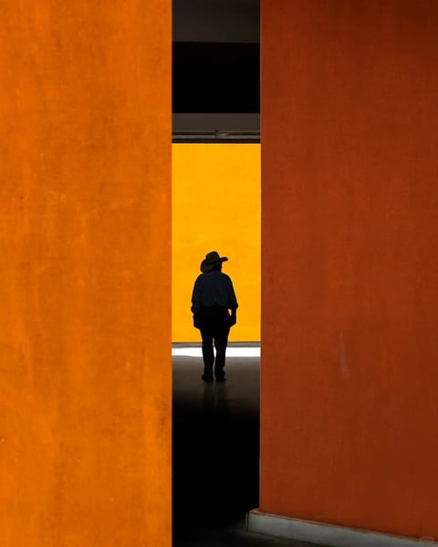 This image is a photorealistic photograph characterized by a bold, minimalist composition with strong geometric framing. The visual aesthetic is clean and graphic, defined by large, flat planes of saturated color. The color language is dominated by a warm, monochromatic palette of deep orange and vibrant yellow, with high contrast between the bright, illuminated background and the deep shadows of the foreground and the silhouetted figure. The lighting approach is directional and dramatic, creating a stark, high-contrast scene where the light source appears to be a bright, even glow from behind the central yellow plane, casting the figure and the foreground architectural elements into near-total silhouette. The texture and material feel are smooth and matte, with the orange walls exhibiting a subtle, uniform grain, while the floor shows a faint, diffuse reflection. The composition is highly structured, using two vertical orange forms to create a narrow, symmetrical frame that directs the viewer's eye to the central figure and the yellow background, establishing a clear sense of depth through layered planes. The shape language is predominantly geometric and rectilinear, with the sharp, clean edges of the architectural forms contrasting with the organic, softened silhouette of the person. The overall mood and atmosphere are contemplative, mysterious, and slightly cinematic, evoking a sense of solitude and focused attention within a stark, modern environment.