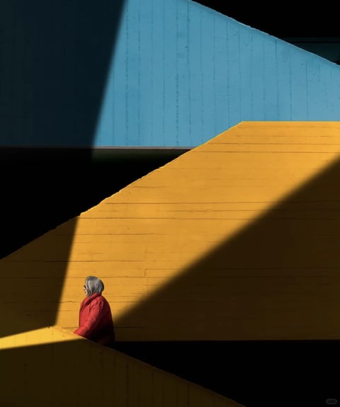 This is a photorealistic architectural photograph characterized by a bold, minimalist aesthetic with strong geometric abstraction. The color language is defined by a high-contrast, saturated palette: a vibrant, sunlit mustard yellow wall dominates the midground, sharply juxtaposed against a deep, cool blue upper wall and areas of pure, inky black shadow. The lighting is harsh and directional, likely from a low-angle sun, casting crisp, angular shadows that slice across the surfaces, creating dramatic diagonal divisions and emphasizing the flat planes. The texture and material feel are rendered with clarity, showcasing the subtle horizontal striations and slight imperfections of painted concrete or plaster, giving a tangible, tactile quality to the large color fields. Compositionally, the image is framed with a strong emphasis on geometric shapes and negative space, using the intersecting planes and the lone human figure to create a sense of scale and depth within a flattened, graphic space. The shape language is almost entirely geometric, defined by sharp, clean edges and straight lines, with the organic form of the person providing a singular point of contrast. The overall mood is one of quiet, contemplative solitude, with a high visual weight and a stark, cinematic atmosphere created by the interplay of light, shadow, and saturated color.