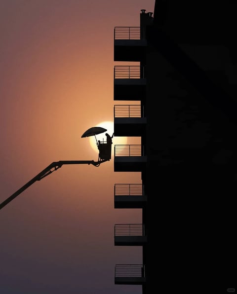 This image is a photorealistic silhouette photograph capturing a moment of human activity against a dramatic sunset. The visual aesthetic is defined by high-contrast backlighting, rendering all foreground elements—the multi-story building, the cherry picker crane, and the worker—as pure, flat black shapes against a luminous sky. The color language is dominated by a warm, monochromatic gradient palette, transitioning from a deep, muted purple at the bottom to a soft, dusty rose and finally to a brilliant, saturated amber-orange where the sun is positioned. The lighting approach is exclusively backlighting from the low, setting sun, which creates a hard, glowing rim around the edges of the silhouettes and bathes the scene in a warm, golden-hour glow. The texture and material feel are smooth and graphic, with the building's balconies and railings presenting sharp, clean geometric lines, while the crane and worker form a more complex, organic silhouette. The composition is vertically oriented, using the strong vertical line of the building on the right to frame the scene, with the crane arm creating a dynamic diagonal leading the eye to the focal point: the worker's basket perfectly aligned with the bright disk of the sun. The shape language is a stark contrast between the rigid, repetitive geometric rectangles of the architecture and the intricate, irregular organic shapes of the machinery and human figure. The mood and atmosphere are serene yet industrious, evoking a sense of quiet, end-of-day labor with a visually weighty, contemplative, and almost cinematic quality.
