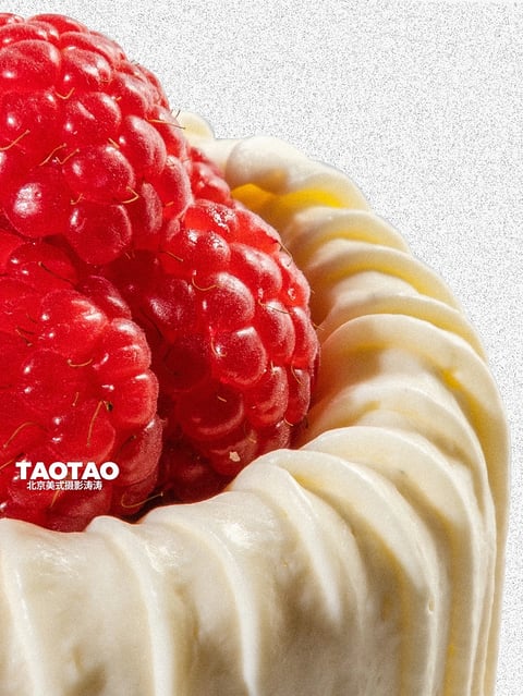 This image presents a hyper-realistic, close-up photographic style with a shallow depth of field, rendering the subject with exceptional detail and clarity. The color language is dominated by a vibrant, saturated crimson red in the raspberries, contrasted against a creamy, off-white base with subtle warm yellow undertones in the shadows, creating a high-contrast and visually striking palette. The lighting is soft and diffused, appearing to come from the upper left, casting gentle highlights on the glossy surfaces of the fruit and the smooth, swirled texture of the base, conveying a fresh and appetizing mood. The texture and material feel are highly tactile, with the raspberries exhibiting a glossy, wet, and bumpy surface with fine, hair-like filaments, while the base appears smooth, creamy, and malleable, like piped frosting or soft cheese. The composition is tightly cropped, focusing on the intersection of the fruit and the base, with the raspberries positioned in the upper left quadrant, creating a dynamic diagonal flow. The shape language is primarily organic, featuring the rounded, clustered forms of the berries and the soft, undulating curves of the base, with sharp, defined edges on the fruit contrasting with the softer, blended edges of the creamy element. The overall mood is indulgent, fresh, and visually rich, emphasizing the luxurious and appetizing qualities of the food.